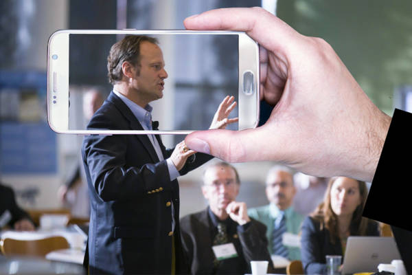 Mann hält im Hintergrund einen Vortrag, im Vordergrund ist eine Hand mit Handy, die auf den Mann hält, sodass dieser auf dem Display erscheint.